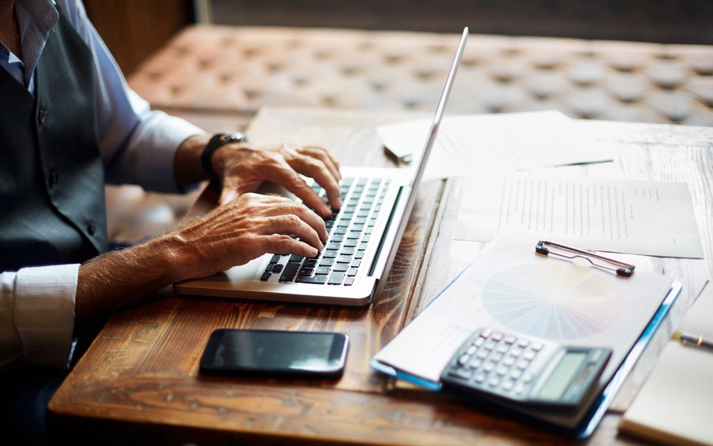 Person working on a laptop with a calculator and clipboard on the desk