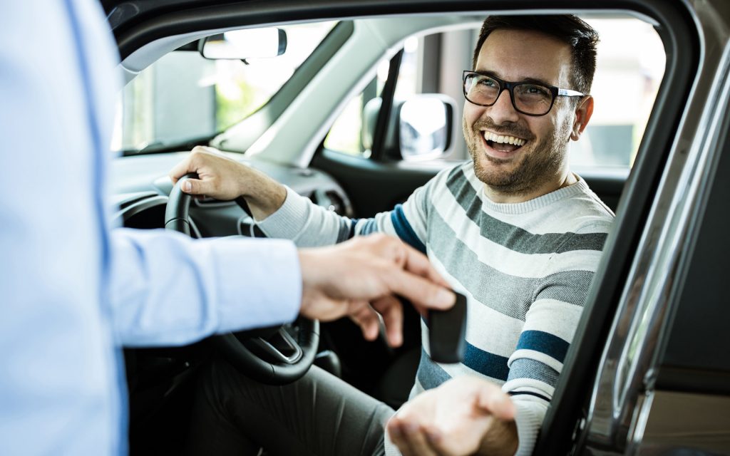 Man smiling behind the wheel of a car as someone hands him the keys
