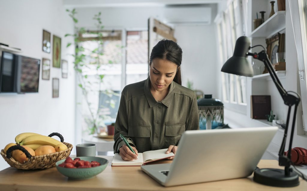 Woman taking notes while working on a laptop in her home office